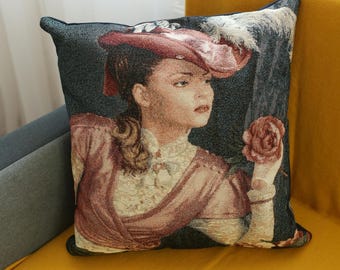 May include: A decorative throw pillow with a tapestry design featuring a woman in a red hat and a rose. The pillow is on a yellow couch.