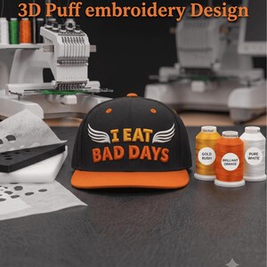 Könnte beinhalten: Eine schwarze Baseballkappe mit orangefarbenem Schirm und dem in 3D-Puff-Stickerei ausgeführten Text I EAT BAD DAYS in Orange mit weißen Flügeln. Die Kappe wird mit drei Stickgarnrollen präsentiert, die als Gold Rush, Brilliant Orange und Pure White beschriftet sind.