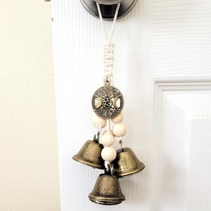 May include: A white macrame door hanger with a triple moon charm and wooden beads. The charm features a pentagram and three crescent moons. The hanger has three brass bells attached to the bottom.