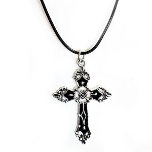 May include: A silver-colored cross pendant necklace with a black cord. The cross has ornate detailing and a dark, possibly black, enamel inlay. The necklace is displayed against a white background.