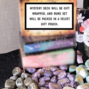 Op de afbeelding: Een verzameling runenstenen en een kaartspel. De tekst op de doos luidt: "MYSTERY DECK WILL BE GIFT WRAPPED, AND RUNE SET WILL BE PACKED IN A VELVET GIFT POUCH." De stenen zijn in verschillende kleuren, waaronder grijs, paars en groen.