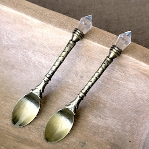 May include: Two antique-style spoons with a bronze finish. Each spoon features a clear crystal at the handle's end and a detailed, twisted design on the stem. The spoon bowls have an oval shape.