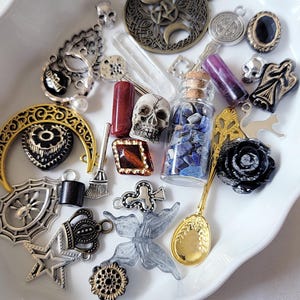 May include: A white bowl filled with a variety of charms, beads, and other small decorative items. The items include a skull, a crescent moon, a butterfly, a spider, a crown, a spoon, a bottle of blue stones, and a black rose.