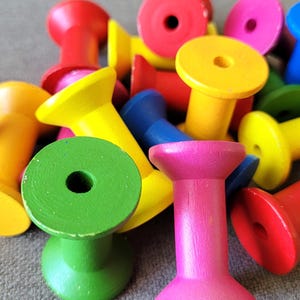 May include: A collection of colorful wooden spools in various shades, including red, yellow, green, pink, and blue. Each spool has a cylindrical body and a flat, circular top with a central hole. The spools are scattered, creating a vibrant and playful arrangement.