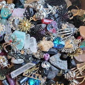 May include: A collection of assorted charms and beads in a variety of colors and materials, including silver, gold, black, and clear. The charms include skulls, spiders, hearts, stars, and crystals. The beads are made of glass, metal, and stone.