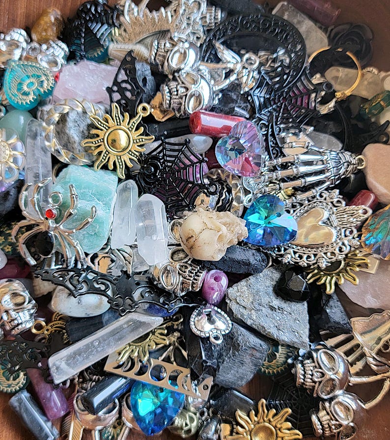 May include: A collection of assorted charms and beads in a variety of colors and materials, including silver, gold, black, and clear. The charms include skulls, spiders, hearts, stars, and crystals. The beads are made of glass, metal, and stone.