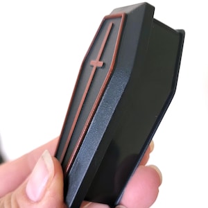 May include: A black coffin-shaped box with a red cross on the front. The box is made of plastic and has a hinged lid.