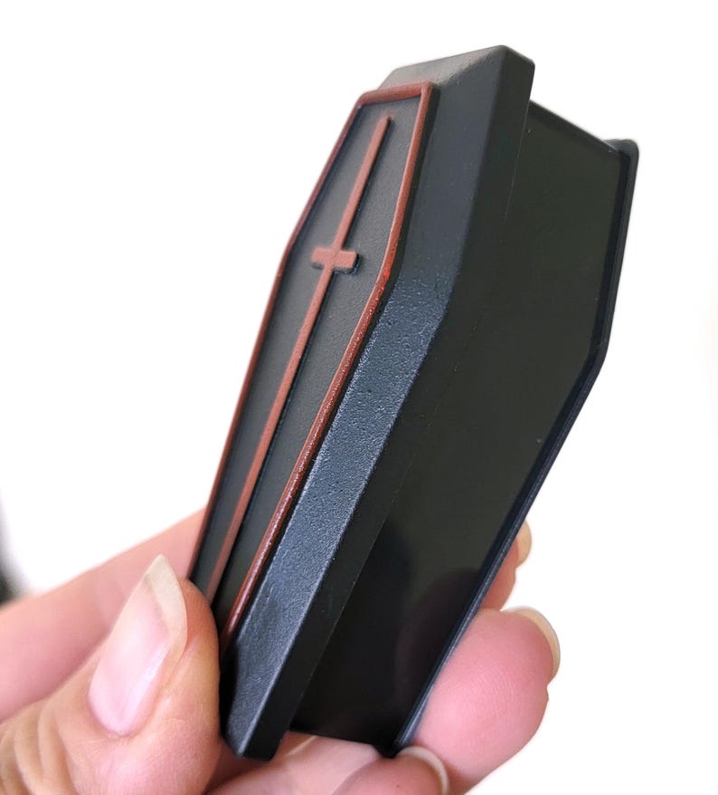 May include: A black coffin-shaped box with a red cross on the front. The box is made of plastic and has a hinged lid.