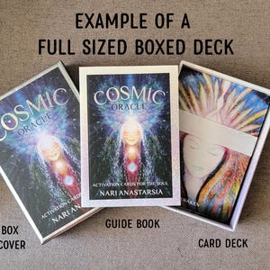Op de afbeelding: Een full-size boxed deck van Cosmic Oracle kaarten. De doos en het gidsboek tonen de titel en illustratie van een vrouw met een gloeiende aura. Het kaartspel is zichtbaar in de doos, met een witte band.