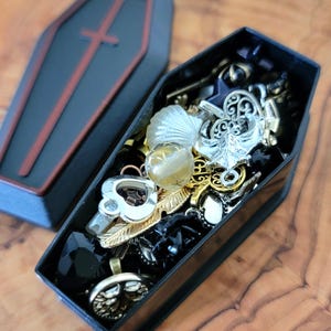 May include: A black coffin-shaped box filled with various charms and trinkets, including a silver anchor, a feather, a heart, and a black stone. The box is open and the charms are visible.