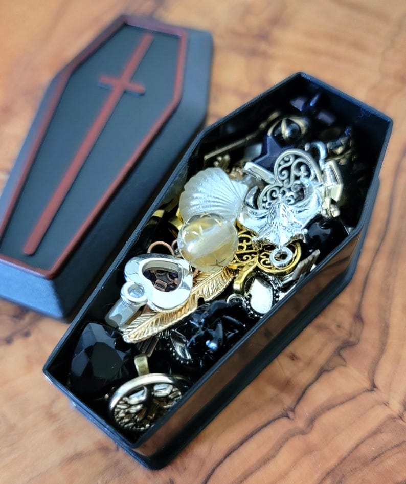 May include: A black coffin-shaped box filled with various charms and trinkets, including a silver anchor, a feather, a heart, and a black stone. The box is open and the charms are visible.