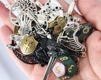 Op de afbeelding: Een verzameling metalen bedels waaronder een zilveren boom, een zwaard, een raaf, een schild met een kruis, een helm, een skelet-eenhoorn en een veelzijdige groen-rode dobbelsteen met gouden cijfers in diverse metalen afwerkingen.