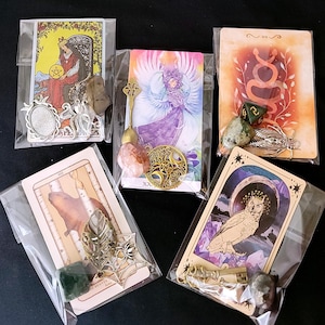 May include: Five tarot cards in clear plastic sleeves, each adorned with unique charms and crystals. Charms include a silver oval frame, an octopus, a spoon, a triple moon pentacle, a cicada, a spider web with a spider, and a key. Crystals vary in color and shape.