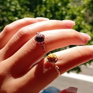 Può includere: Due anelli fatti a mano con filo metallico. Un anello presenta una pietra strutturata marrone scuro, mentre l'altro ha una pietra giallo-marrone. Gli anelli sono ambientati su uno sfondo verde sfocato, con l'attenzione focalizzata sui gioielli.