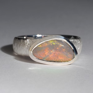 May include: A silver ring with a textured band and a large oval opal gemstone. The opal has a milky white base with flashes of green, orange, and red.