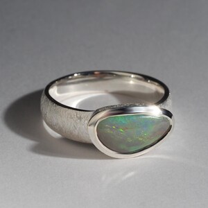 May include: A silver ring with a textured band and a bezel-set oval opal gemstone. The opal has a green, blue, and white iridescent sheen.