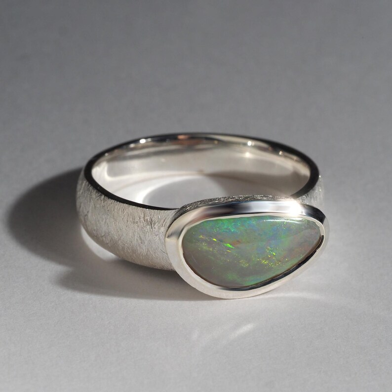 May include: A silver ring with a textured band and a bezel-set oval opal gemstone. The opal has a green, blue, and white iridescent sheen.