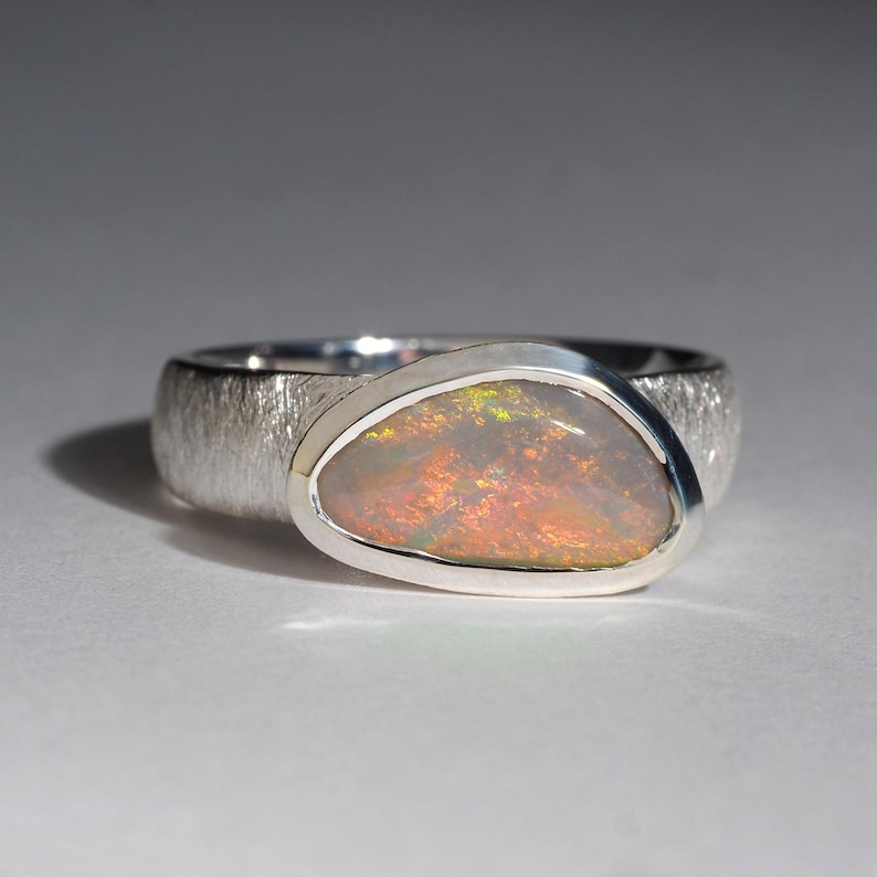 May include: A silver ring with a textured band and a large oval opal gemstone. The opal has a milky white base with flashes of green, orange, and red.