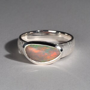 May include: A silver ring with a textured band and a large oval opal gemstone. The opal has a rainbow of colors, including red, orange, yellow, green, blue, and purple.
