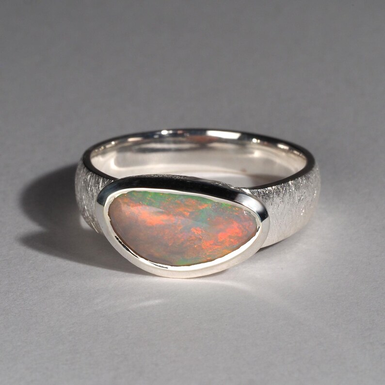 May include: A silver ring with a textured band and a large oval opal gemstone. The opal has a rainbow of colors, including red, orange, yellow, green, blue, and purple.