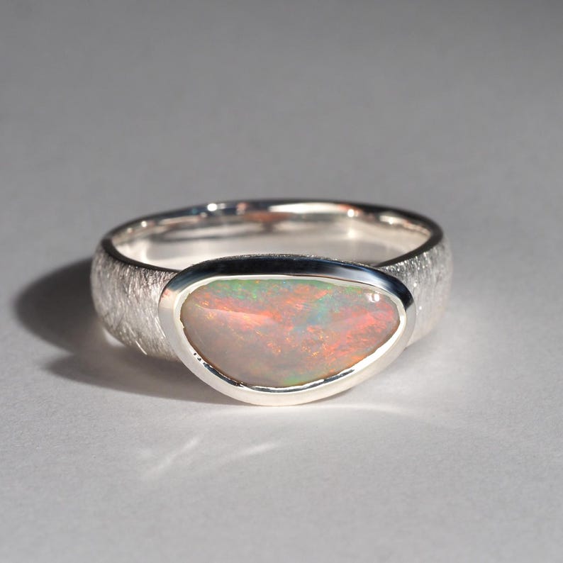 May include: A silver ring with a textured band and a large, oval-shaped opal gemstone. The opal has a milky white base with flashes of pink, green, and blue.