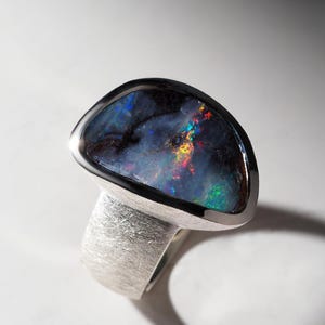 Könnte beinhalten: Silberring mit einem dunklen Opal-Edelstein, der schillernde Farben zeigt. Der Opal ist in einer polierten Silberfassung gefasst, und der Ring hat eine strukturierte, gebürstete Oberfläche. Ein auffälliges Schmuckstück.