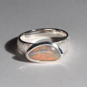 May include: A silver ring with a textured band and a large oval opal gemstone. The opal has a milky white base with flashes of orange, red, and green.