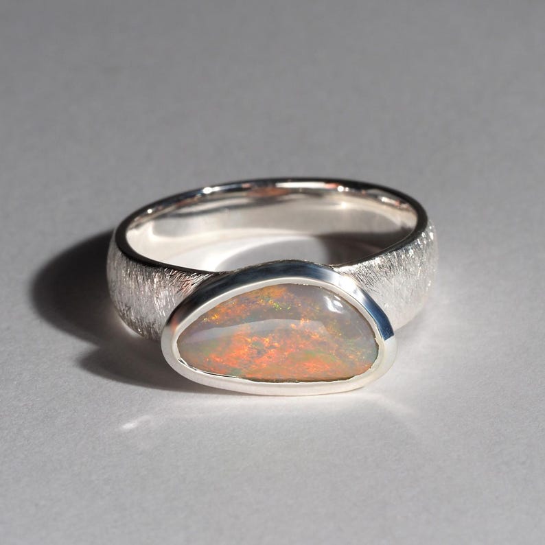 May include: A silver ring with a textured band and a large oval opal gemstone. The opal has a milky white base with flashes of orange, red, and green.