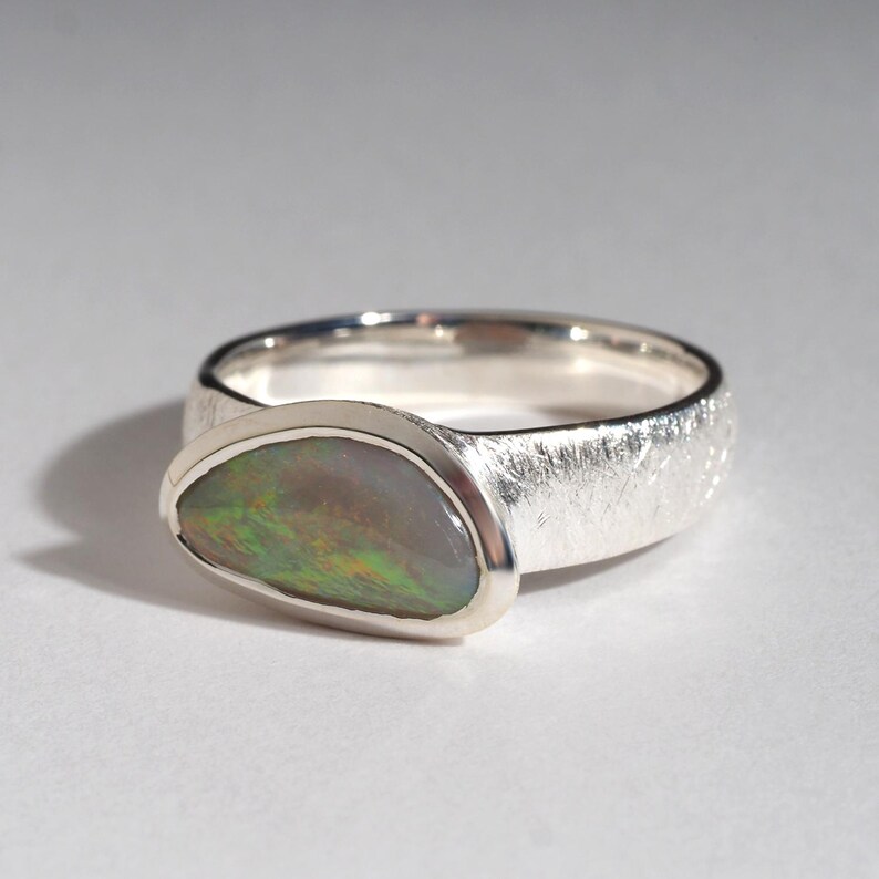 May include: A silver ring with a textured band and a large, oval-shaped opal gemstone. The opal has a green, blue, and brown iridescent sheen.