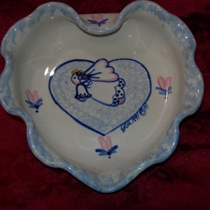 May include: A white heart-shaped ceramic dish with a blue and white angel design and the text "Erick Ray 1991". The dish has a scalloped edge and is decorated with pink flowers.