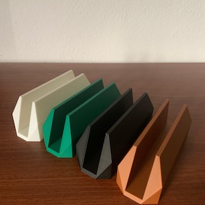 Può includere: Cinque supporti per telefono di diversi colori realizzati in plastica. I supporti sono disposti in fila da sinistra a destra, con il supporto bianco a sinistra e il supporto marrone a destra. Tutti i supporti hanno la stessa forma, con un'apertura a forma di V nella parte anteriore.