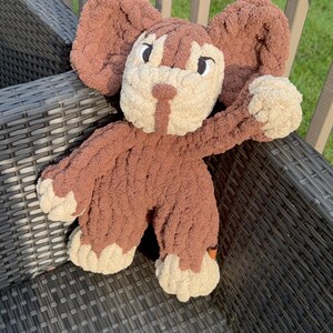 May include: A handmade brown and cream-colored plush toy in the shape of a bunny. The bunny has large floppy ears, a cream-colored face, paws, and a body made of soft, textured yarn. The toy is sitting on a gray wicker chair.