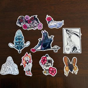 Puede incluir: Una colección de nueve pegatinas de vinilo con ilustraciones inspiradas en el anime. Los diseños incluyen personajes con acentos florales, una figura azul de múltiples caras, una criatura oscura con un bastón, un panel de manga y una polilla naranja.