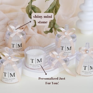 May include: Set of five clear glass candle jars with lids, each decorated with a small white bow and a single shiny stone. The jars contain white candles with personalized labels that read "T/M Taylor & Matthew August 7, 2022".