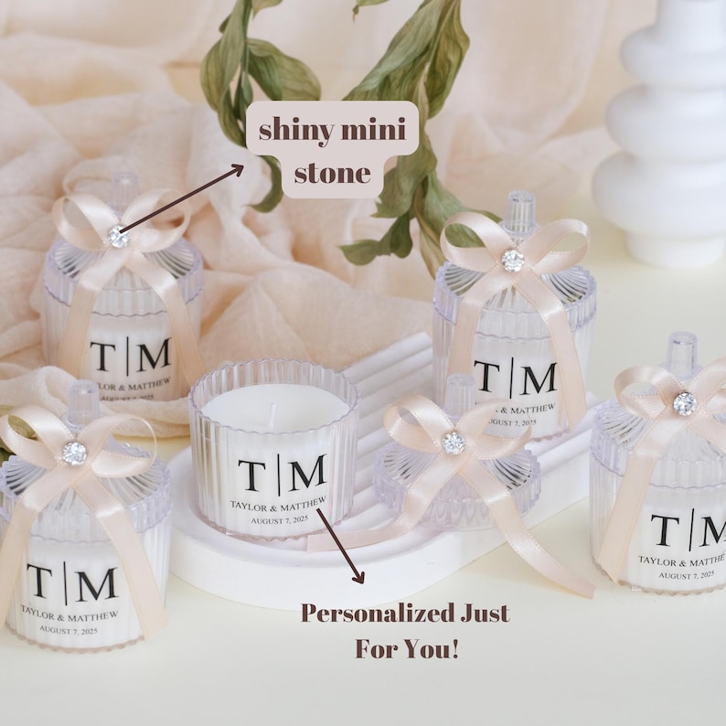 May include: Set of five clear glass candle jars with lids, each decorated with a small white bow and a single shiny stone. The jars contain white candles with personalized labels that read "T/M Taylor & Matthew August 7, 2022".