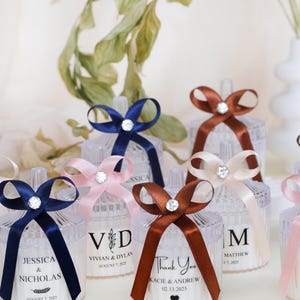 May include: Assortment of clear glass candle jars with personalized labels and different colored ribbons. The labels include names and dates, such as "Jessica & Nicholas, August 7, 2025" and "Vivian & Dylan, August 7, 2025".
