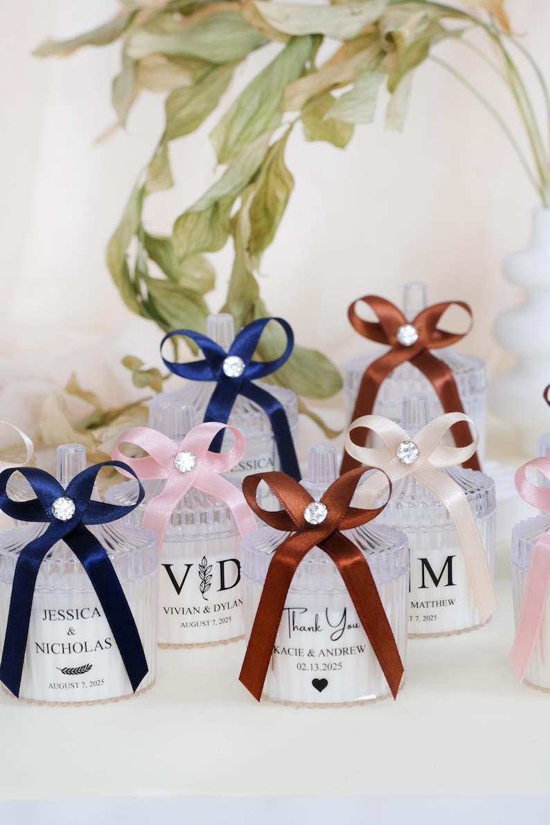 May include: Assortment of clear glass candle jars with personalized labels and different colored ribbons. The labels include names and dates, such as "Jessica & Nicholas, August 7, 2025" and "Vivian & Dylan, August 7, 2025".