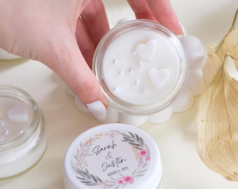 May include: Small glass jars with white lids and white wax inside. The lids are decorated with floral designs and the text "Sarah & Justin August 5, 2023".
