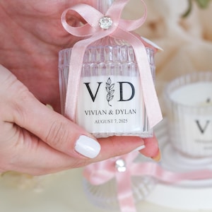 May include: A clear glass candle jar with a pink ribbon and a rhinestone bow. The jar has a label that says "V&D Vivian & Dylan August 7, 2025".