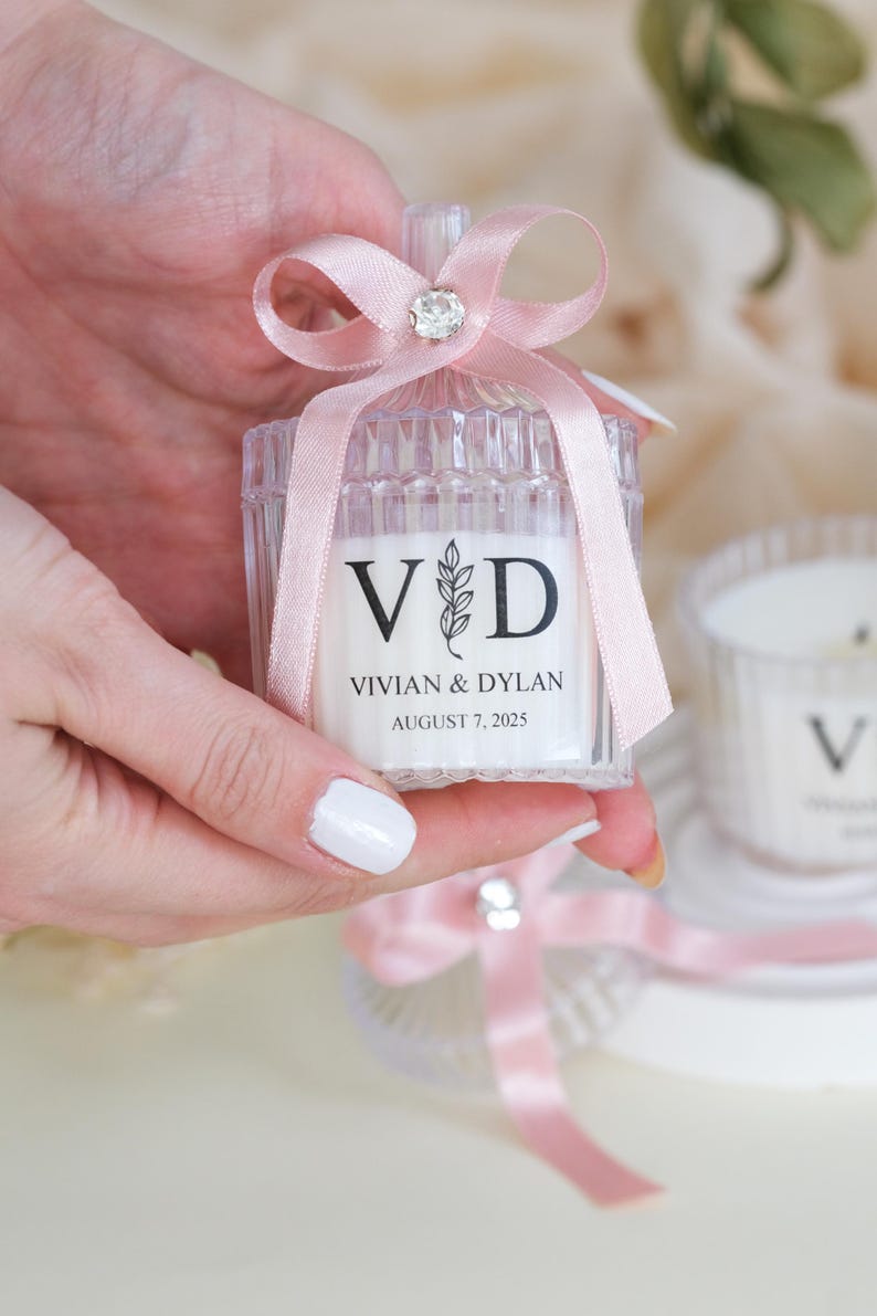May include: A clear glass candle jar with a pink ribbon and a rhinestone bow. The jar has a label that says "V&D Vivian & Dylan August 7, 2025".