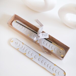 Könnte beinhalten: Ein hellgrauer Räucherstäbchenhalter mit Mondphasen-Design und Goldblatt-Akzenten. Der Halter befindet sich in einer rechteckigen Schachtel mit einem Band und einem Etikett mit der Aufschrift "HEATHER & JASON". Ein zweiter Halter befindet sich neben der Schachtel. Weiße Dekorationsgegenstände im Hintergrund.