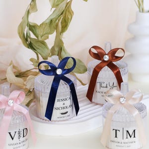 May include: Four clear favor boxes with different colored ribbons and personalized labels. The labels include the names Jessica & Nicholas, Vivian & Dylan, Kacie & and Taylor & Matthew, along with their wedding dates.