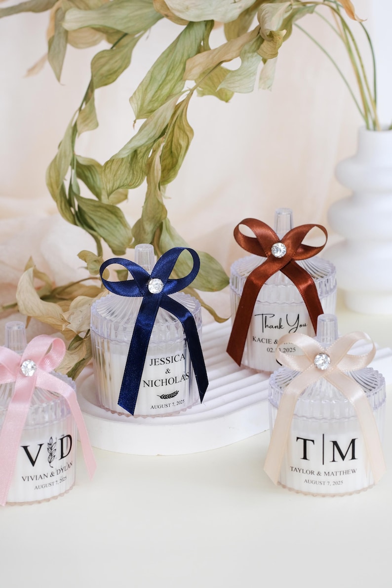May include: Four clear favor boxes with different colored ribbons and personalized labels. The labels include the names Jessica & Nicholas, Vivian & Dylan, Kacie & and Taylor & Matthew, along with their wedding dates.
