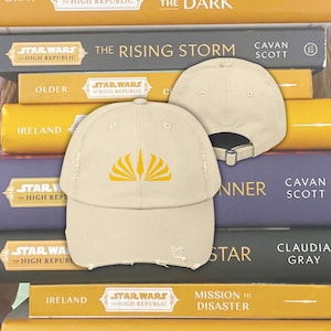 Könnte beinhalten: Eine beige, abgenutzte Baseballkappe mit einem gelben gestickten Logo einer stilisierten Sonnenexplosion. Das Logo stammt aus der Buchreihe Star Wars: The High Republic.