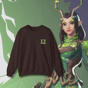 Könnte beinhalten: Ein dunkelbraunes Sweatshirt mit einem kleinen, neongrünen Emblem auf der linken Brust. Das Sweatshirt wird vor einem grünen und violetten Hintergrund mit einer Charakterillustration präsentiert. Es ist ein lässiger Stil.