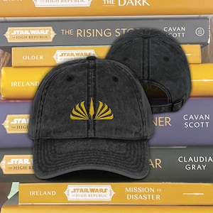May include: Black baseball cap with a yellow embroidered design. The cap has a curved brim and an adjustable strap. The cap is displayed in front of a stack of books with the words "Star Wars" and "High Republic" on the spines.