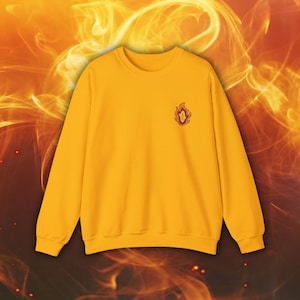 Könnte beinhalten: Ein gelber Pullover mit einem kleinen gestickten Flammenmotiv auf der linken Brust.