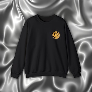 Könnte beinhalten: Schwarzer Pullover mit einem goldenen gestickten Design auf der linken Brust. Das Design zeigt einen Kreis mit einem stilisierten Buchstaben "O" darin.