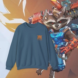 Peut inclure: Un sweat-shirt ras du cou bleu sarcelle avec un motif orange sur la poitrine. Le motif est un emblème stylisé avec du texte en dessous. L'arrière-plan présente un personnage de dessin animé avec une arme.