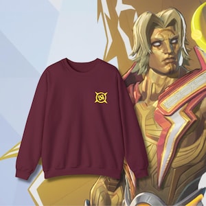 Puede incluir: Una sudadera burdeos de cuello redondo con un pequeño emblema amarillo en el pecho. La sudadera se muestra frente a una ilustración dorada de un personaje con cabello rubio y un atuendo rojo y dorado.